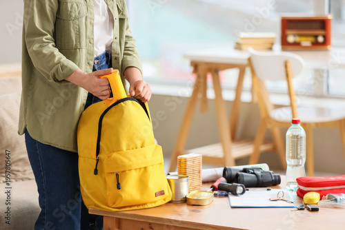 Photos Woman packing emergency supplies into yellow backpack at home, preparing disaste