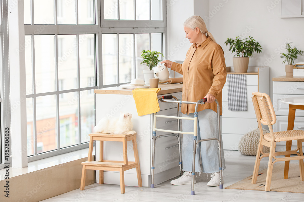 Fototapeta premium Mature woman with walker and cute cat pouring tea in kitchen