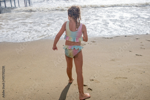 Фотография carefree child, Little girl in pastel swimsuit running barefoot towards foamy oc