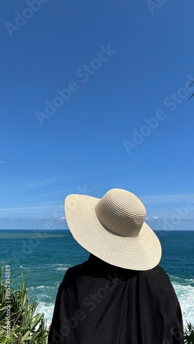 Muslim woman wearing black outfit and beach hat looking at ocean. Travel lifestyle concept with wide copy space. Vertical 