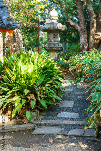 Traditional Japanese garden path with stone lantern, lush green foliage, and stepping stones. Peaceful Zen landscape design with cast iron plants and bamboo under natural sunlight.