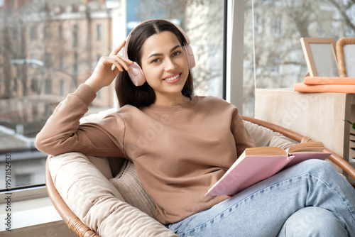 Beautiful young woman in headphones listening to audiobook with book and sitt...