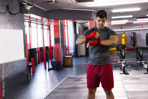 Mid adult man wrapping red hand wraps around wrists at gym with heavy bags and whiteboard