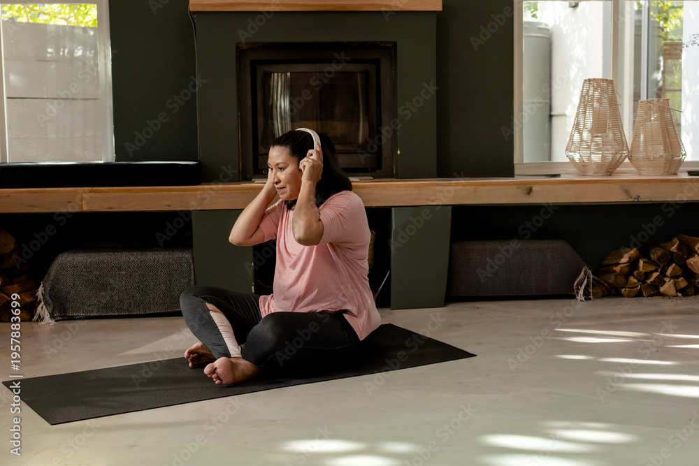 Naklejka premium Asian woman sitting cross-legged on black yoga mat in home living area holding over-ear headphones