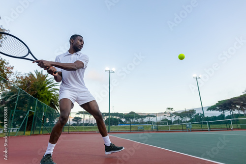 Fotografia African-American male preparing two-handed backhand on court in white holding ra