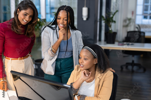Diverse female colleagues collaborating around computer monitor in open office with potted plants