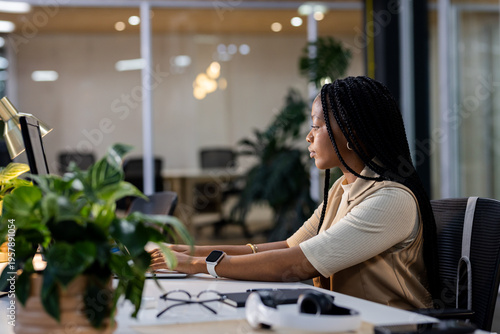 Office desk monitor and keyboard glowing under warm lamp near plant, eyeglasses by glass walls