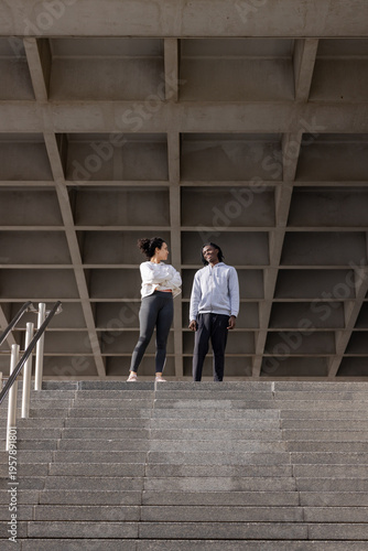 Diverse female friends in activewear standing atop concrete stairway under coffered ceiling