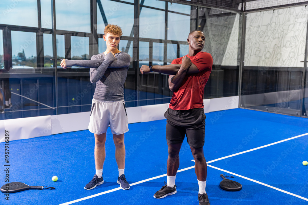 Obraz premium Diverse male partners stretching arms on rooftop blue padel court with rackets, balls, smartwatches