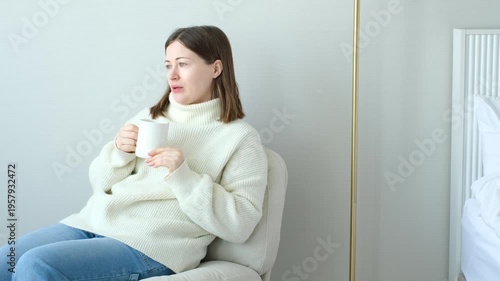 Young woman sits on a chair and drinks coffee from a white mug.