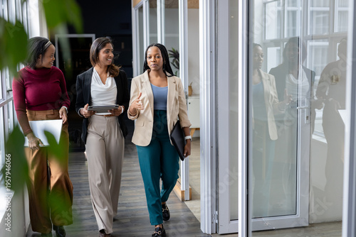 Diverse female colleagues in business attire walking office corridor with laptop, tablet and folder