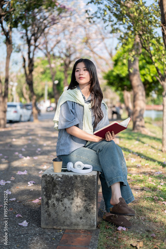 Foto Pretty asian woman looking away from a book while sitting cross legged on concre