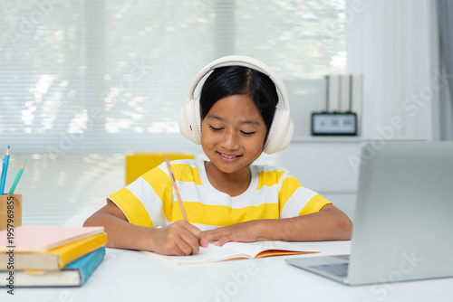 Asian Girl with Headphones Taking Notes While Learning Online on Laptop at Home