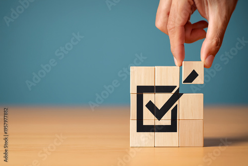 Completion and approval concept. Abstract background with hand placing final wooden block into cube puzzle forming black check mark inside square symbolizing success, validation, and task achievement.