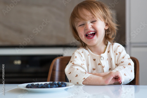 A two-year-old cute girl is sitting at the dinner table eating blueberries or bilberries. Baby food. Feeding a child. Healthy snack for children.