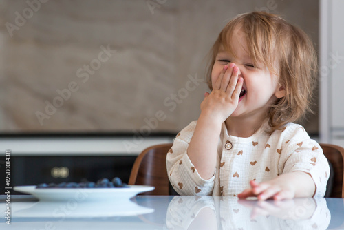 A two-year-old cute girl is sitting at the dinner table eating blueberries or bilberries. Baby food. Feeding a child. Healthy snack for children.