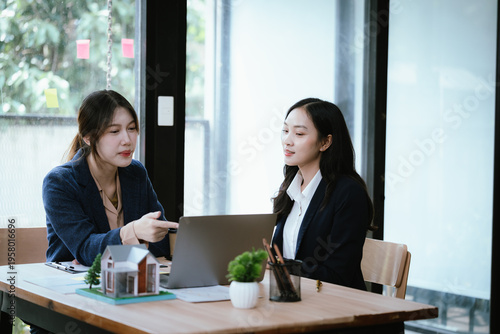 Two businesswomen discussing a house model and documents at a desk near a window, representing real estate consultation, property investment, and professional teamwork.