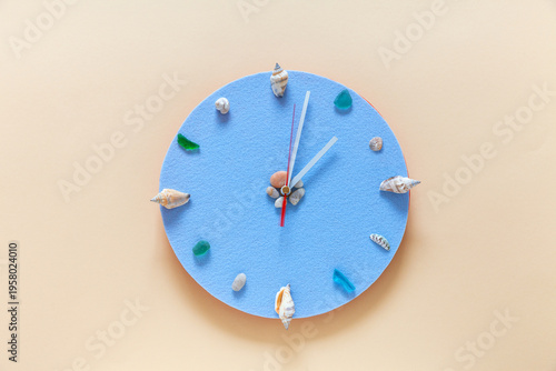 Time for a seaside holidays. Handmade clock with a light blue dial and shells and pebbles instead of numbers on light-colored wall. Summer vacation concept