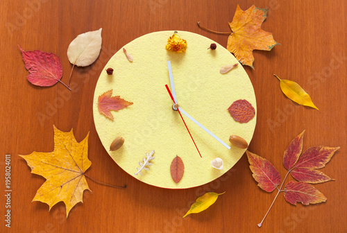 Time of autumn. Handmade clock with a yellow dial, with fallen leaves instead of numbers, on a wooden background. Concept for the arrival of autumn season