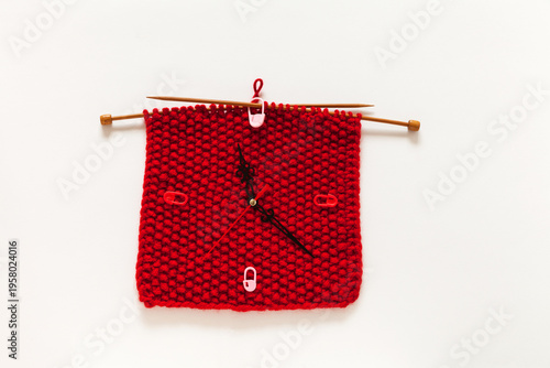 Time for crafting and knitting. Handmade wall clock with a red knitted square dial on wooden knitting needles on a white background. Hobby and craft concept