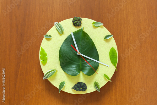 Time of spring. A handmade clock with a yellow dial, decorated with green young leaves instead of numbers, on a wooden background. Concept for the arrival of spring
