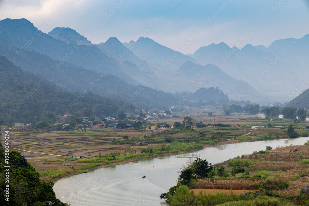 Naklejka premium A scenic view of Song Mien River flowing through a valley in Quan Ba, Ha Giang, Vietnam, with mountains in the background and a small boat carrying people on the water near Cao Bang in Vietnam.