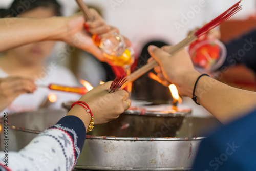 Multiple hands hold incense bundles and candles over a metal oil lamp, sharing a moment of collective prayer and offering.