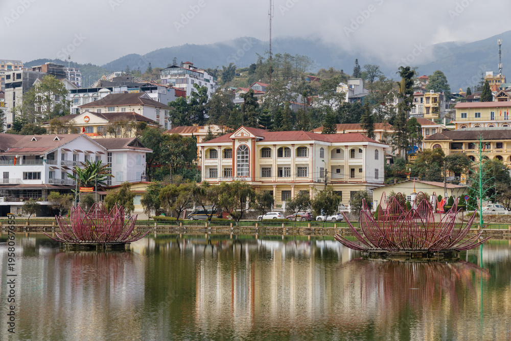 Fototapeta premium Scenic view of Sa Pa Lake with lotus sculptures, reflecting the town's architecture, in Vietnam. A tranquil scene showcasing local art and urban landscape.