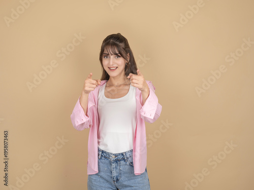 Tableau sur toile Smiling young woman pointing at camera with both hands, wearing pink shirt and denim, standing against beige studio background with copy space