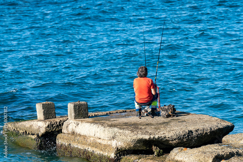 Solo Fisherman Sitting on Concrete Pier Fishing Rods Over Blue Ocean Water on a Sunny Day