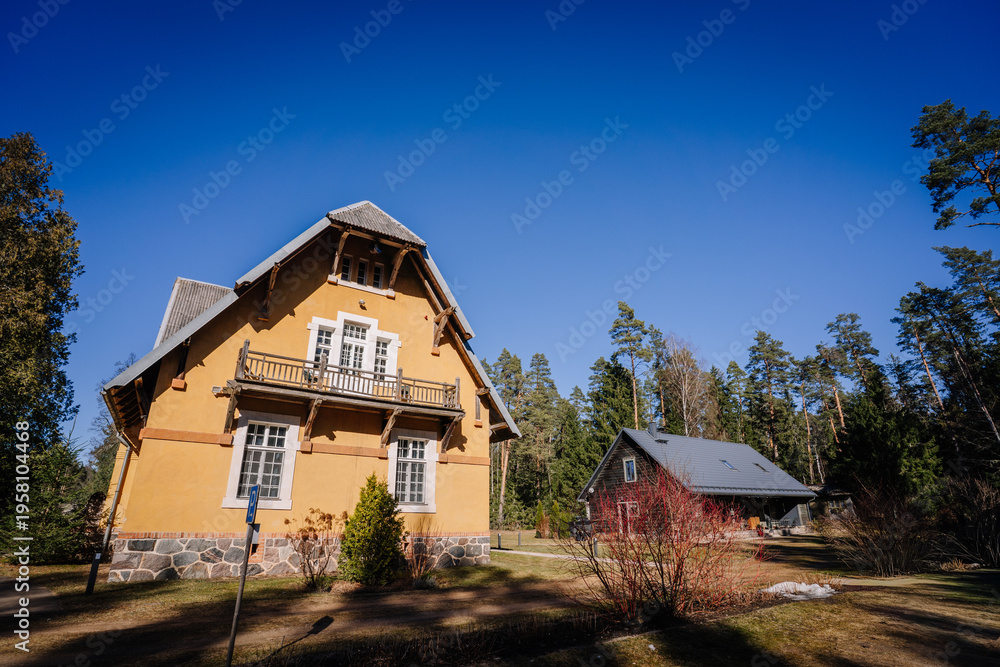 Obraz premium Two-story yellow house with balcony and gray roof surrounded by trees and grass under a clear blue sky in a rural setting