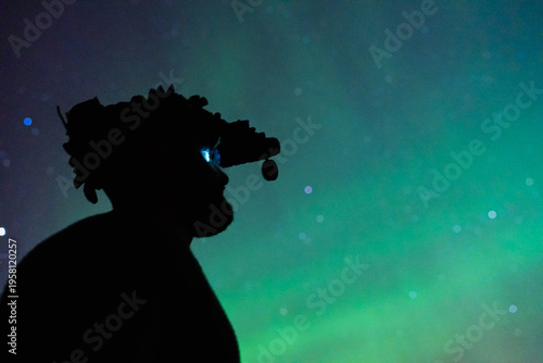 Tactical silhouette of a man in night vision goggles observing vibrant green Aurora Borealis in Greenland during night operation