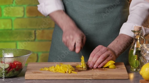 A male chef slices yellow bell pepper with a knife on a cutting board to prepare a salad.