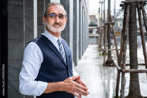 Confident mature Caucasian businessman standing sideways with hands clasped gesture wearing formal suit outdoors in modern city