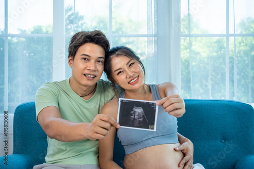 Joyful Asian couple sharing baby ultrasound, tender pregnancy moment at home, looking at camera