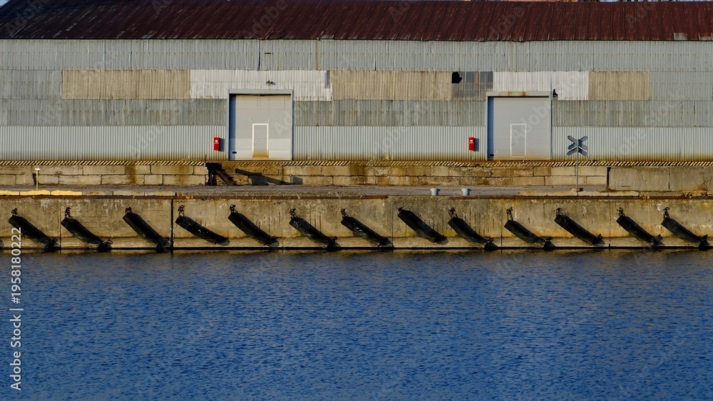 Naklejka premium Industrial harbor pier with black fenders and rhythmic long shadows on concrete wall. Corrugated metal warehouse background. Maritime logistics and port infrastructure. Sunset lighting.