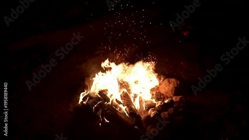 A warm orange fire with dancing yellow flames and red heat burning wood inside a fireplace creates a glowing light against the dark night background