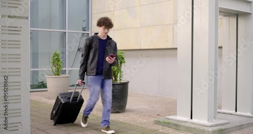 Entering glass walkway, man pulling black suitcase, checking phone for time, showing to other man