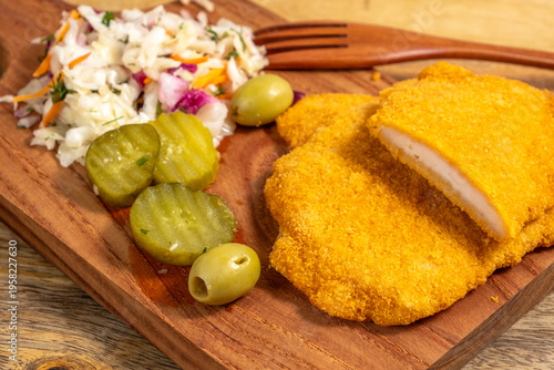 Food background with crispy breaded chicken cutlet, pickled olives and cucumbers on wooden platter.
