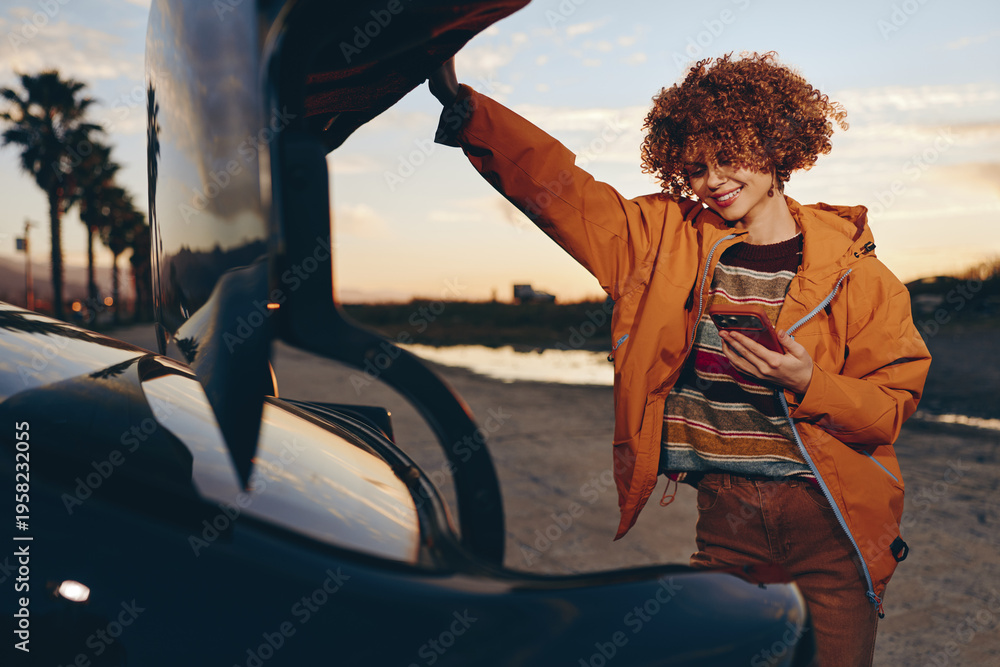 Fototapeta premium Smiling woman with curly hair wearing a rainbow sweater and orange jacket stands by a car at sunset holding a smartphone, enjoying a relaxed lifestyle moment outdoors.