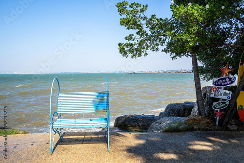 Blue metal bench facing a windy sea under a clear sky, with a tree, decorative signs and surfboard on the side, creating a relaxing seaside atmosphere.