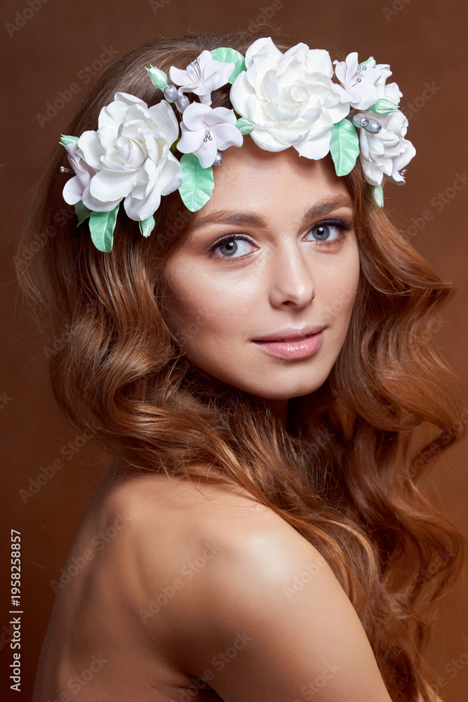 Fototapeta premium Close-up of a beautiful portrait of a young girl with a flower wreath in her hair. She is wearing a delicate flower crown with green leaves. The concept of beauty, spa, manicure, and skin care