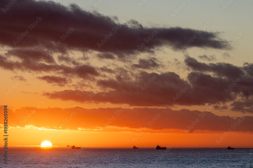 Fototapeta premium Sunset over the Atlantic ocean with freight ships passing near the descending sun, near the port of Huelva, Spain