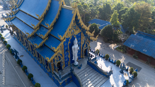 Aerial view of the resplendent Wat Rong Suea Ten Temple, where sapphire hues meet golden accents under the watchful gaze of a pristine white Buddha statue, Chiang Rai, Thailand.