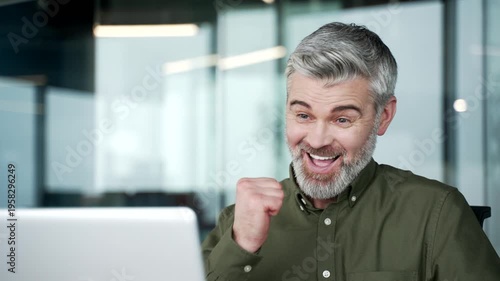 Close-up of gray-haired businessman smiling after reading positive news on laptop. Man reacting with joy and relief while checking good message at workplace in modern office, cheerful and inspired.
