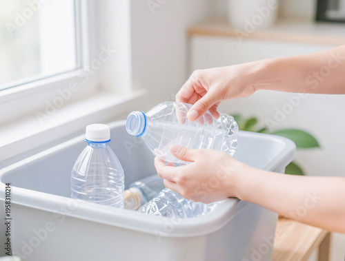 Close-up of a person's hands placing an empty clear plastic bottle into a grey recycling bin indoors. Bright minimalist setting near a window, emphasizing ecology and sustainable waste management