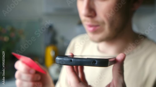 Naive man holding bank card and smartphone, dictating his financial data to scammer during phone call at home, becoming victim of online fraud, identity theft. Concept of phone fraud or scammer.