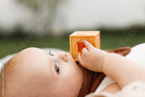 Beautiful happy baby lying on blanket and playing with toys on background of sunny summer garden. Childhood. Portrait of cute infant relaxing in park, enjoying summer outdoors