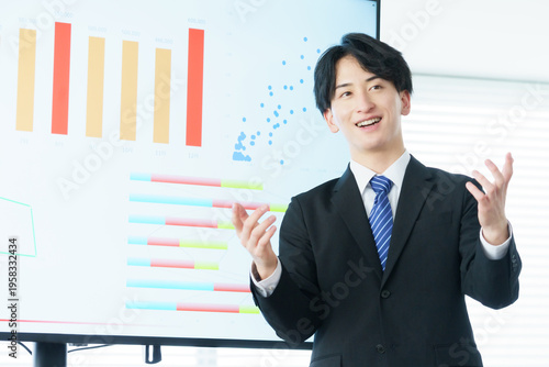 オフィスでディスプレイに映ったグラフを説明しながらプレゼンをする若い日本人男性