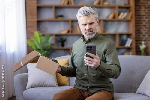 Mature man holding an open cardboard box and smartphone, looking disappointed and stressed after receiving an incorrect or damaged online order at home
